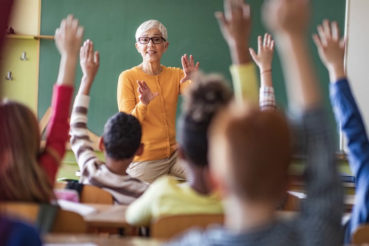 A teacher stands in front of a class full of children with their hands raised