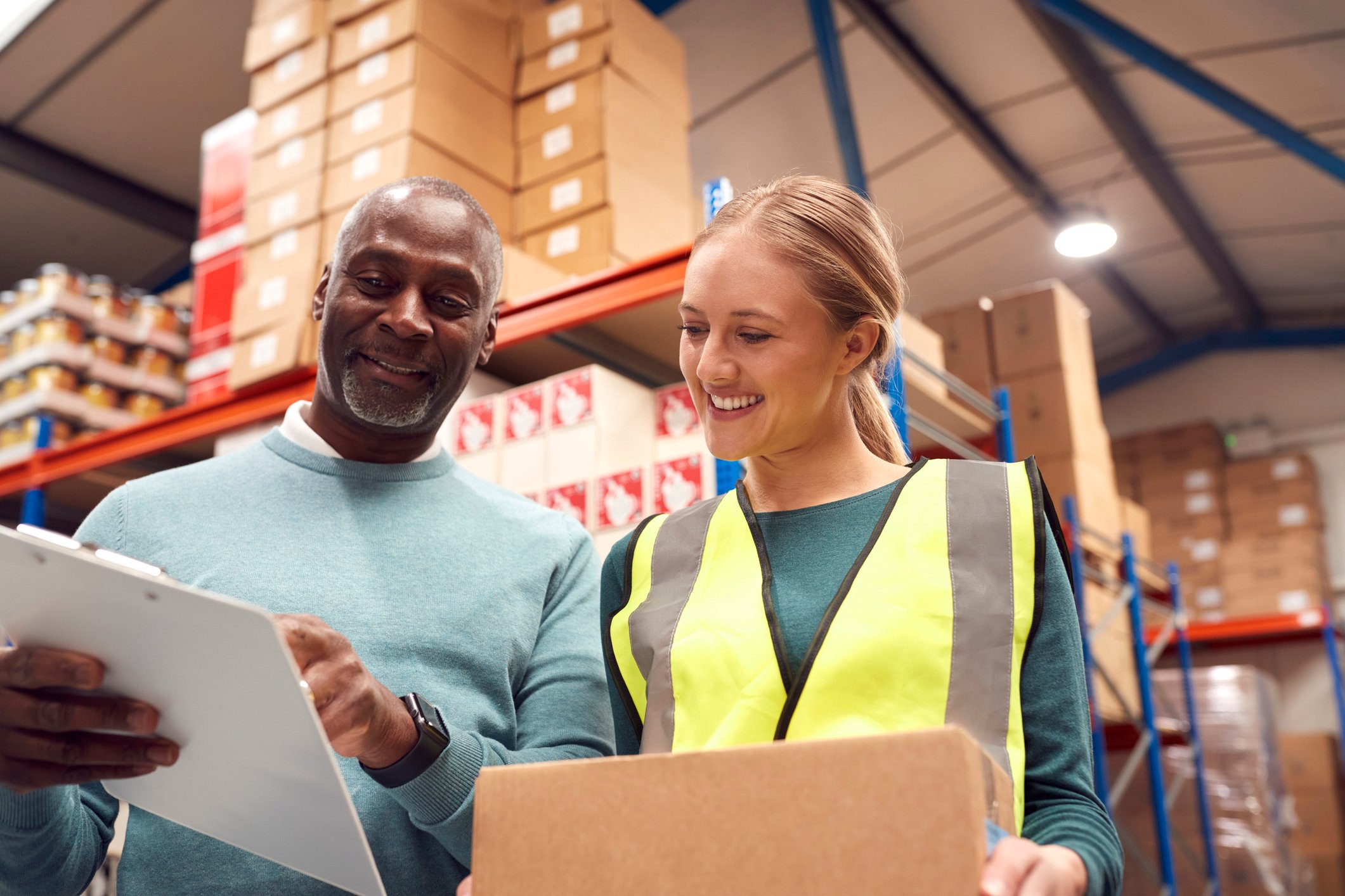 A man and woman smile as they look at information on a clipboard in a warehouse area.